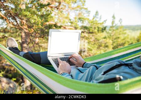 A man working on a laptop while lying in a hammock in the woods. Empty screen. Self-isolation, freelancing, remote work and distancing. Top view Stock Photo