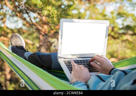 A man working on a laptop while lying in a hammock in the woods. Empty screen. Self-isolation, freelancing, remote work and distancing. Top view Stock Photo