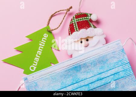 Tag with text and toy Santa Claus with medical mask on a pink background, top view. Concept on the topic of quarantine for the New Year holidays. Stock Photo