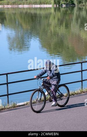 An older man rides his bike wearing a face covering bandana. In a park in Flushing, Queens, New York. Stock Photo