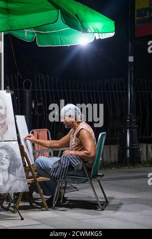 Sudak, Russia - September 15, 2020: An artist of mature age in a headscarf draws portraits on a city street in the evening under a lantern. Stock Photo