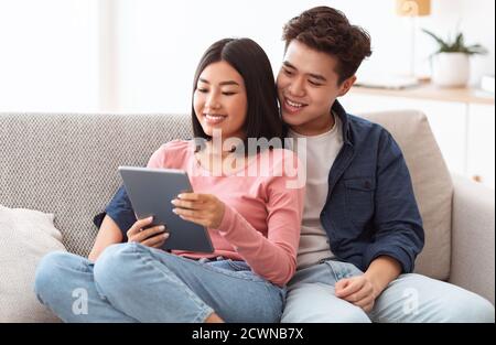 Cheerful Asian Couple Using Digital Tablet Sitting On Couch Indoors Stock Photo