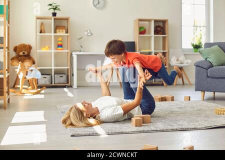 Happy young mother lying on carpet, holding her son on legs in raised hands and having fun at home