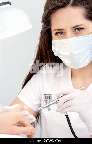 Portrait of manicure master uses airbrush wearing mask in nail salon Stock Photo