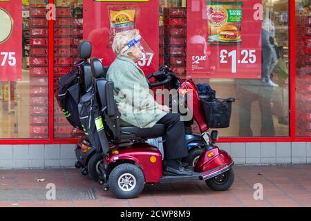 An elderly lady wearing a face shield sitting on a mobility scooter outside a supermarket during the coronavirus pandemic Stock Photo