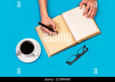 Woman writing in a diary with a cup of coffee on a blue background in a top view Stock Photo