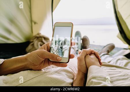 Close-up of unrecognizable couple of hikers relaxing in tent and taking photo of crossed legs against lake Stock Photo