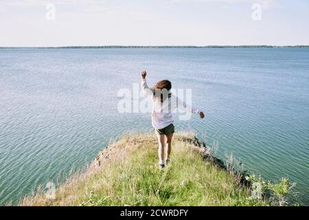 Rear view of carefree curly-haired girl in shirt running along cliff covered with grass to lake and swinging arms
