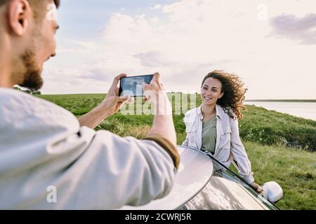 Over shoulder view of bearded man using smartphone while photographing beautiful girlfriend with wind in her hair against field and lake Stock Photo