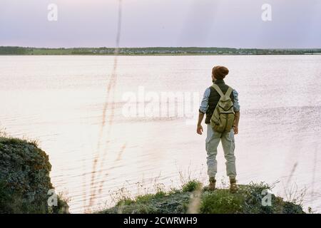Rear view of young male backpacker in hat standing on edge of cliff and contemplating lake while enjoying hike Stock Photo