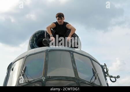 A young guy in black and unmarked in military boots sitting on the roof of a helicopter. Stock Photo