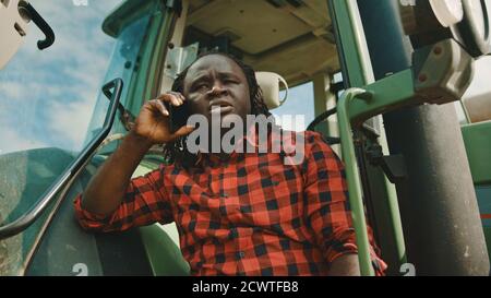 Young african farmer speaking on the smartphone while leaning on the green tractor. High quality photo Stock Photo