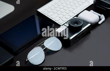 office table, with retro camera, sunglasses, headphone charger, white keyboard, partial view waterproof case and silver computer base, on black backgr Stock Photo