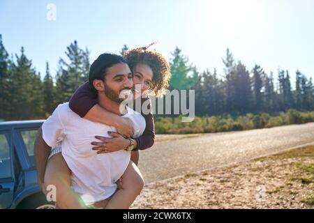 Happy affectionate couple piggybacking at sunny summer roadside Stock Photo
