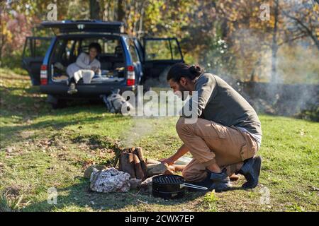 Man preparing campfire outside car in sunny autumn field Stock Photo