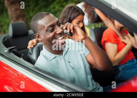 Happy man talking on smart phone in convertible with family Stock Photo