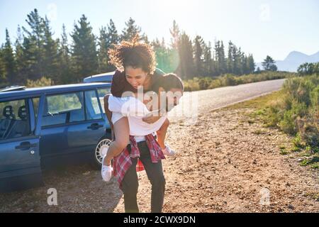 Playful young couple piggybacking outside car at sunny remote roadside Stock Photo