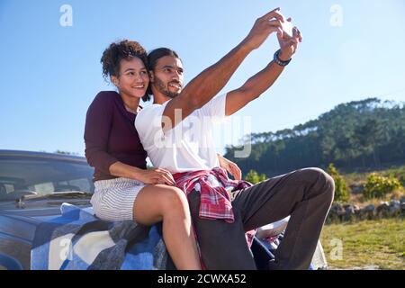 Happy young couple taking selfie on hood of car at sunny roadside Stock Photo
