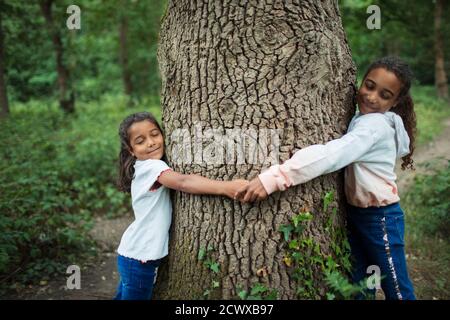 Serene sisters hugging tree trunk in woods Stock Photo