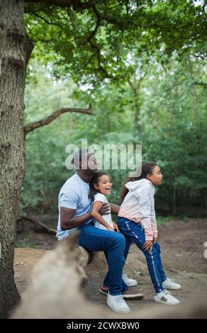 Happy father and daughters sitting on fallen log in woods Stock Photo