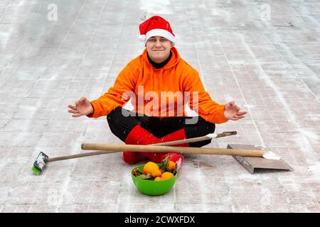 A man in a red Christmas hat and boots, in a orange jacket, sits in a lotus position on the site of his yard with a brush and a shovel. He cleaned the snow from the backyard and was pleased with it. Stock Photo