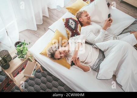 Top view of the couple lying in cozy bed in bedroom and browsing internet and checking mails using each other modern smartphone. Modern technology, so Stock Photo