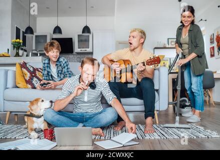 Quarantine time: Family at living room together. Father distance working using laptop, mother cleaning the room, older son playing acoustic guitar and Stock Photo