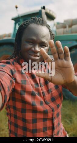 Video call. African farmer waving to the smartphone camere in front of the tractor. High quality photo Stock Photo