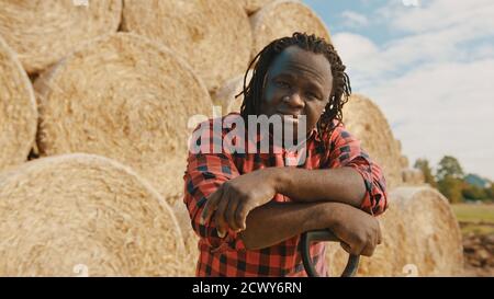 Young african farmer resting on the pitchfork handle. Hay roll stack in the background. High quality photo Stock Photo