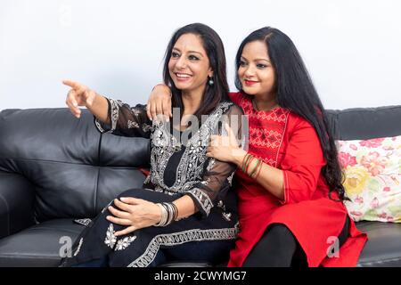 Two smiling female friends sitting on a couch at home and having a good time, laughing out happily, pointing towards something. Stock Photo