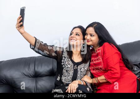 Two smiling female friends sitting on a couch at home and taking a selfie with phone. Stock Photo