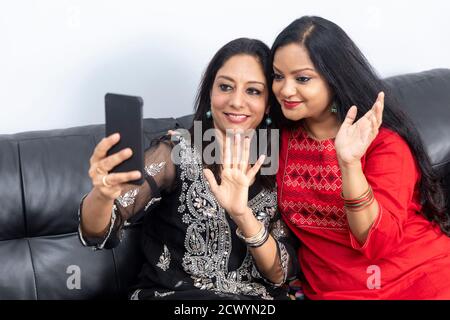 Two smiling female friends sitting on a couch at home and taking a selfie with phone, video conference with friend, waving hands. Stock Photo