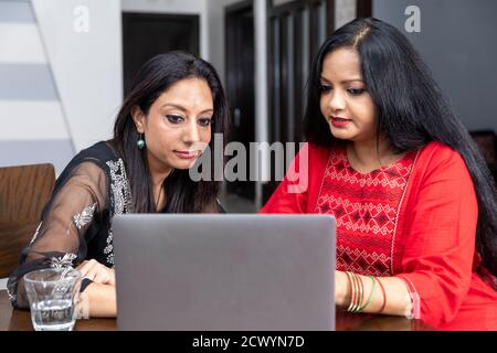 Two female friends sitting at home with laptop infront of them, smiling and looking into camera, video call with friend. Stock Photo