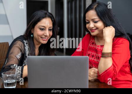 Two female friends sitting at home with laptop infront of them, smiling and looking into camera, video call with friend. Stock Photo