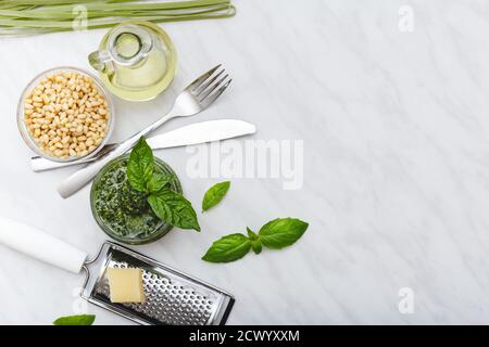 Pesto sauce with ingredients: pine nuts, basil, olive oil, parmesan, cutlery, Raw green fettuccine on white marble table. Recipe making Pesto Stock Photo