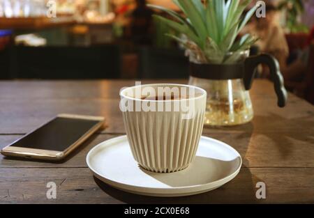 Cup of Hot Coffee on a Wooden Table with Blurry Smartphone and Potted Plant Stock Photo