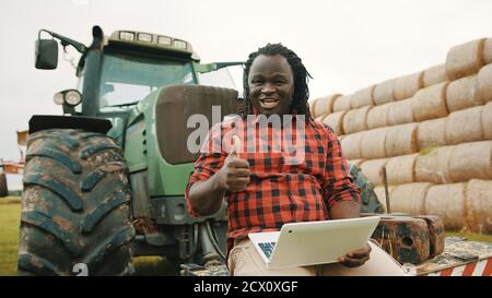 Young african farmer using tablet while sitting on green tractor. Haystack in the background. High quality photo Stock Photo