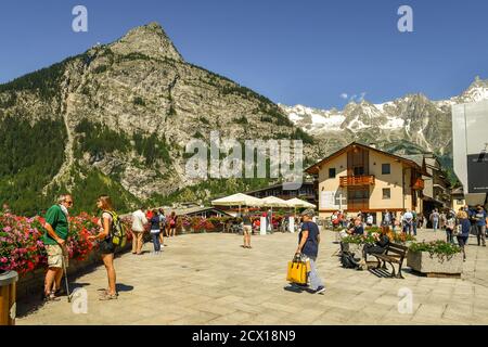 Piazza Abbé Henry, the main square of the famous Alpine town, with tourists and backpackers, Mont Chetif and Mont Blanc massif in summer, Courmayeur Stock Photo