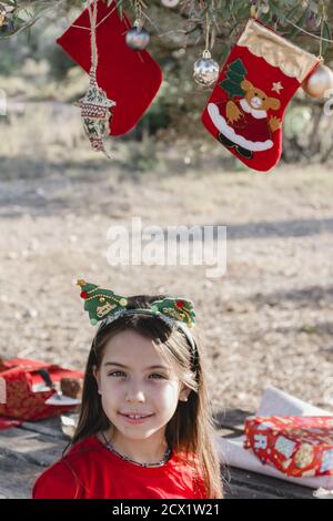 A girl dressed in red and with a Christmas headband under the Christmas decorations hanging from an olive tree in the field after a picnic Stock Photo