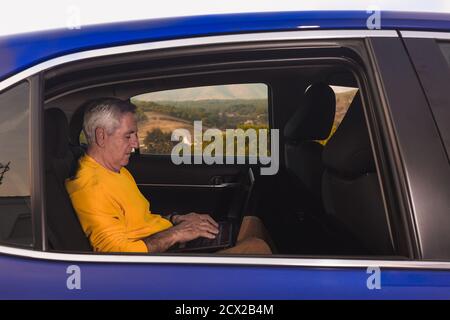Side view of aged male traveler sitting on back seat and typing on laptop keyboard during road trip in countryside Stock Photo