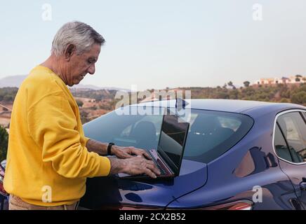 Side view of an elderly traveler sailing with a laptop in the trunk of a modern blue vehicle in the countryside. Stock Photo