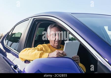 Elderly man in yellow sweatshirt sitting on passenger seat of car and using tablet during road trip Stock Photo