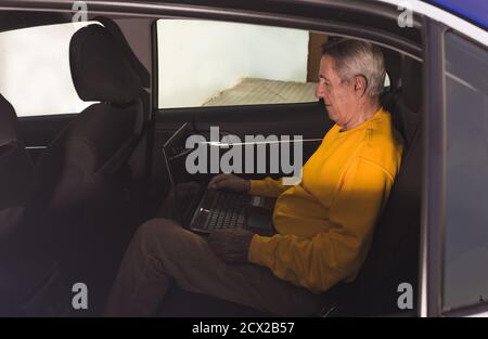 Side view of aged male traveler sitting on back seat and typing on laptop keyboard during road trip in countryside Stock Photo