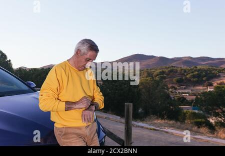 Senior man in casual clothes leaning on modern vehicle and checking wristwatch during road trip in countryside Stock Photo
