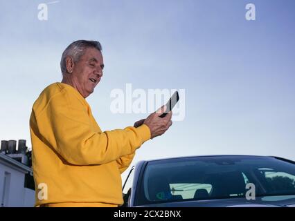 Side view of the smiling old man in a yellow sweatshirt navigating the cell phone GPS map while standing near a modern vehicle during a road trip Stock Photo