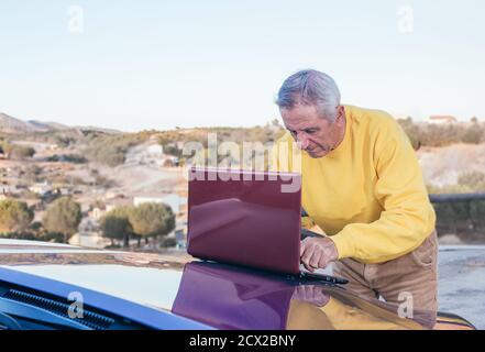 Front view of elderly male traveler browsing laptop on hood of electric blue modern vehicle in countryside Stock Photo