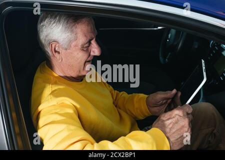 Side view of a smiling older man in a yellow sweatshirt sitting in the passenger seat of a car and wearing a pill during the trip. Stock Photo