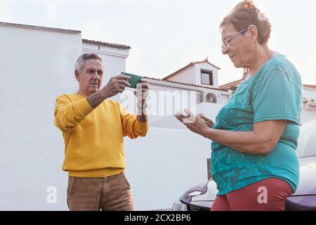 Elderly man and woman browsing mobile phones while standing near car on street of town during road trip Stock Photo