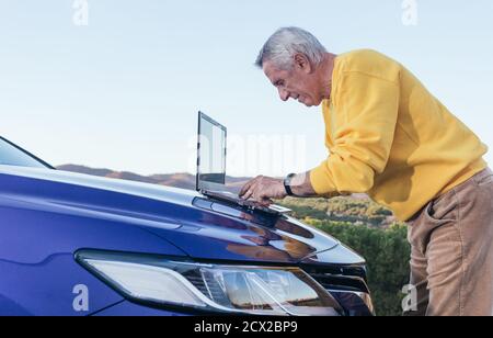 Side view of elderly male traveler browsing laptop on hood of blue modern vehicle in countryside Stock Photo