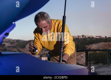 Elderly traveler with flashlight opening the trunk of the vehicle looking for a fault in a blue electric car Stock Photo